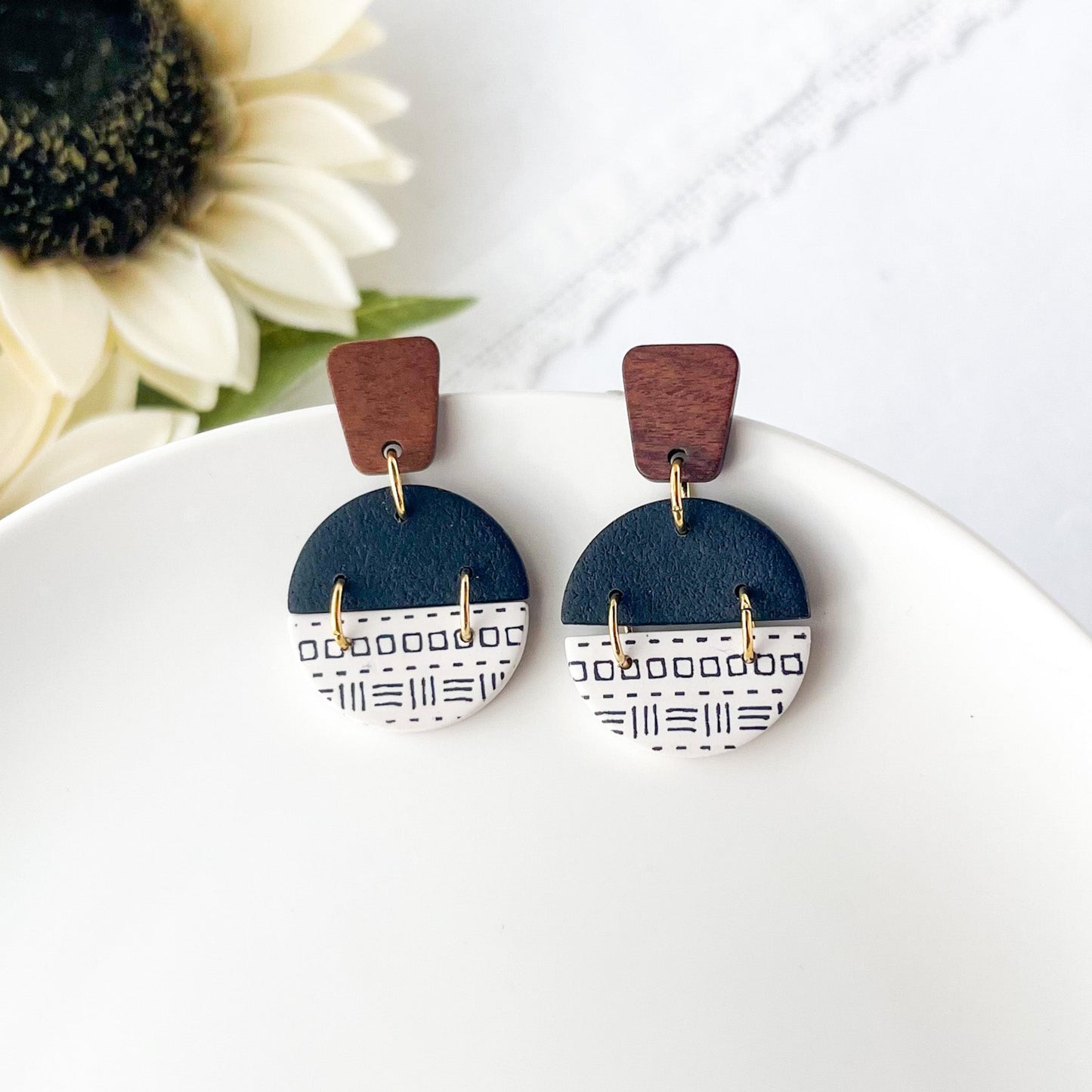 Pair of earrings with wooden tops and black and white printed polymer clay on a white surface with sunflower in the background