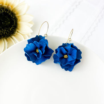 Pair of indigo blue large flower polymer clay statement earrings dangling from gold hoop lying on a white plate with floral background. The blue flowers have multiple textured petals and has a gold colored stamen.