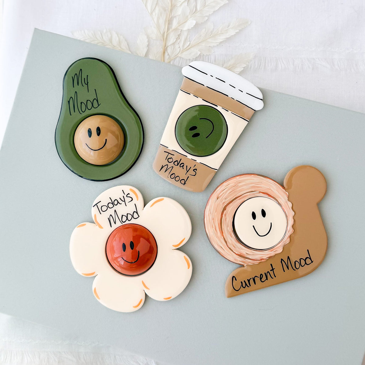 Four handmade polymer clay mood magnets shaped like an avocado, coffee cup, flower, and snail, each with a swappable happy face