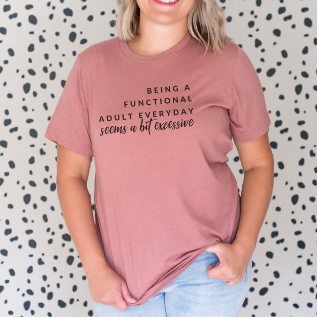 Person wearing a mauve t-shirt with text that reads being a functional adult everyday seems a bit excessive, against a patterned background