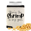 A beige can cooler with the phrase “You’re the Shrimp to My Grits,” standing on a white background with peanuts scattered in front of the can cooler.