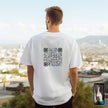 Person wearing a white t-shirt with a QR code design, standing against an outdoor cityscape.