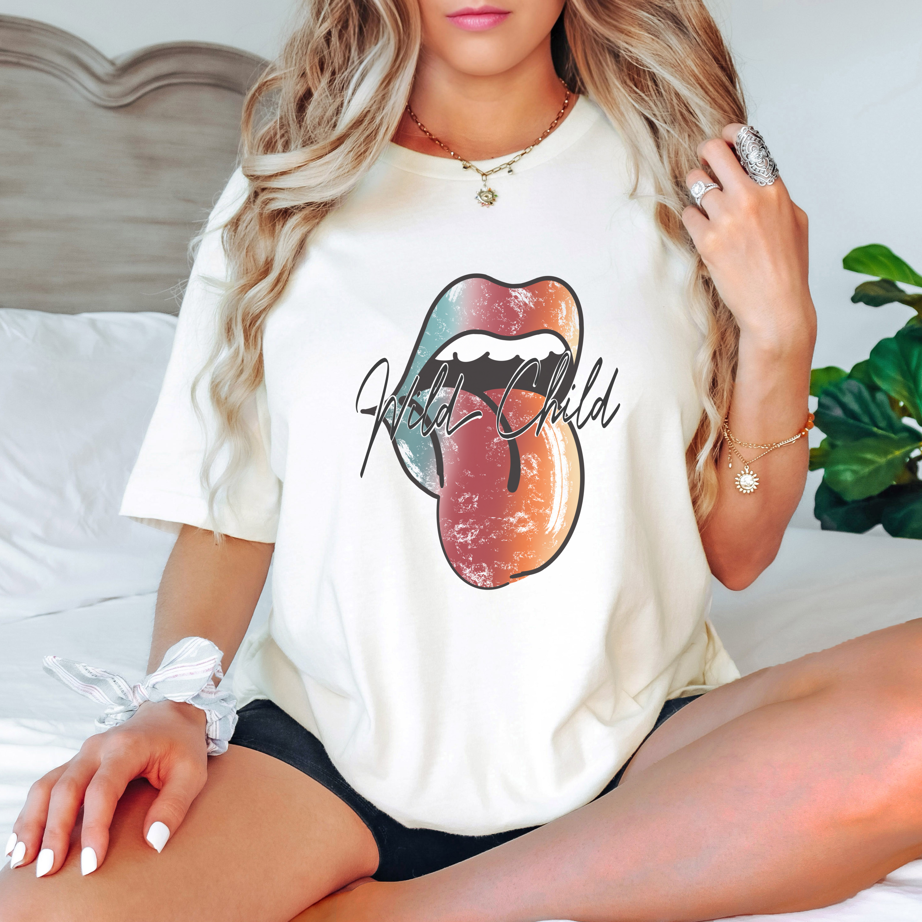 Woman sitting on a bed wearing a white t-shirt with a colorful graphic design of a tongue with text 