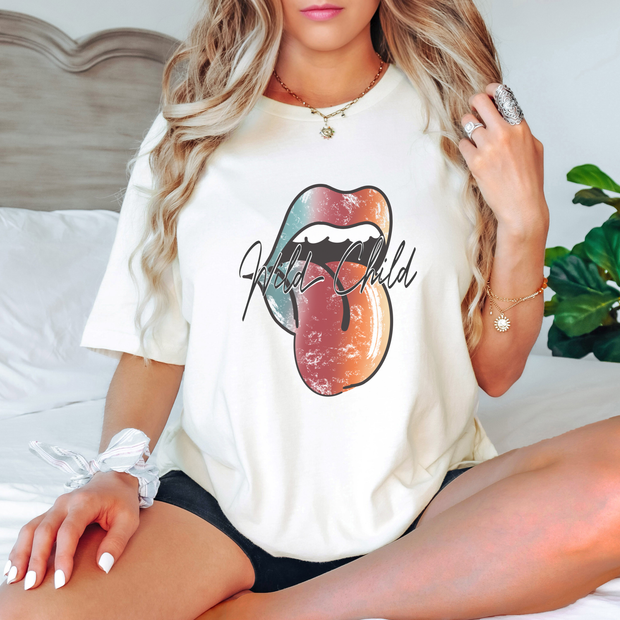 Woman sitting on a bed wearing a white t-shirt with a colorful graphic design of a tongue with text "Wild Child".