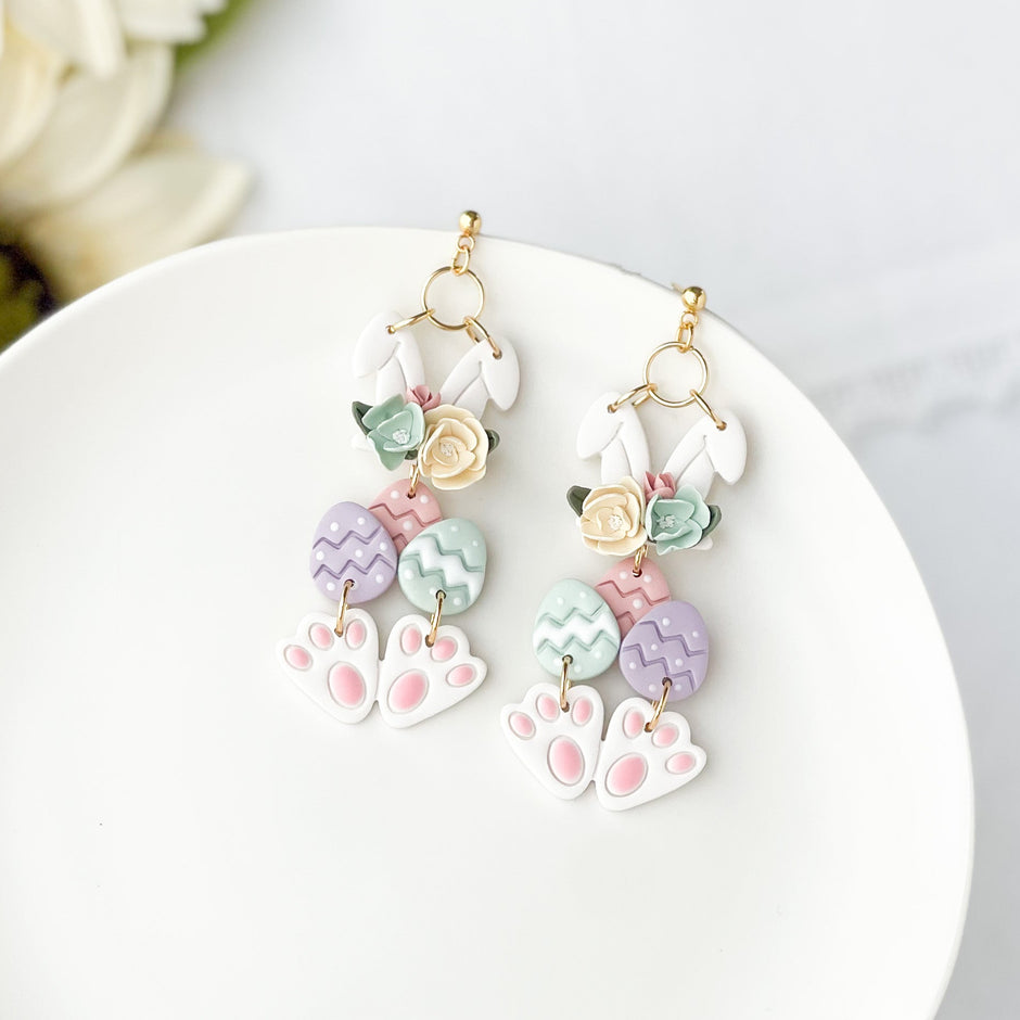 A pair of floral patterned earrings featuring an Easter bunny design, with clip-on closures, displayed on a white plate.