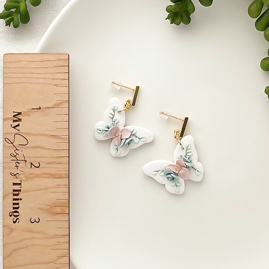 Butterfly earrings on a wooden stand with a white dish and greenery in the background
