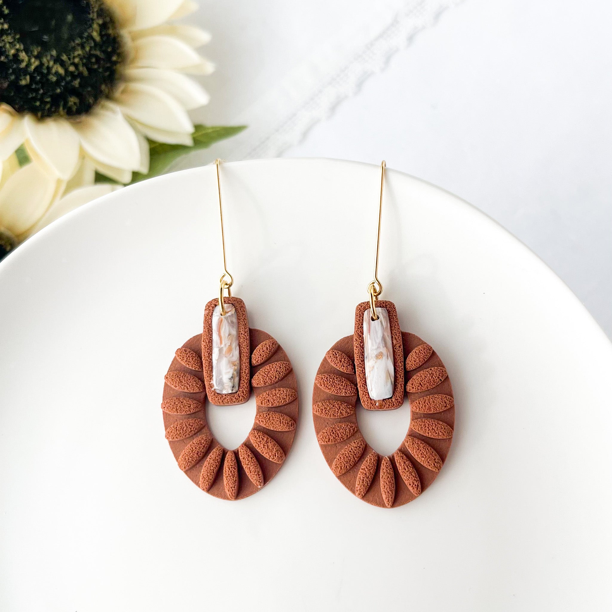 Brown textured earrings with white stones on a white plate with flowers in the background