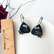 Pair of black earrings with gold designs on a white surface with a wooden ruler for scale.