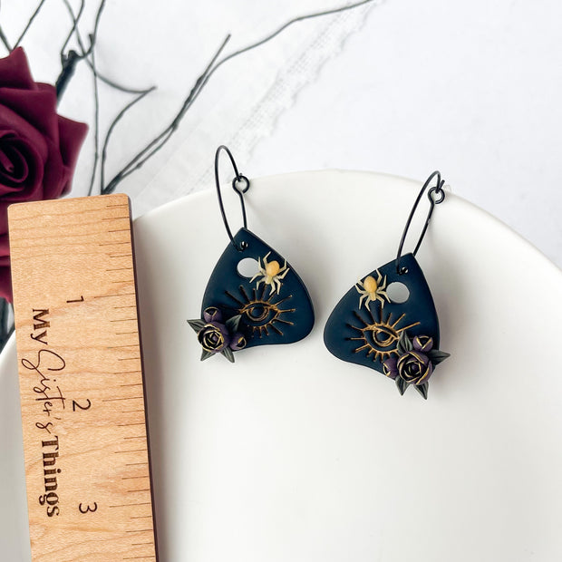 Pair of black earrings with gold designs on a white surface with a wooden ruler for scale.