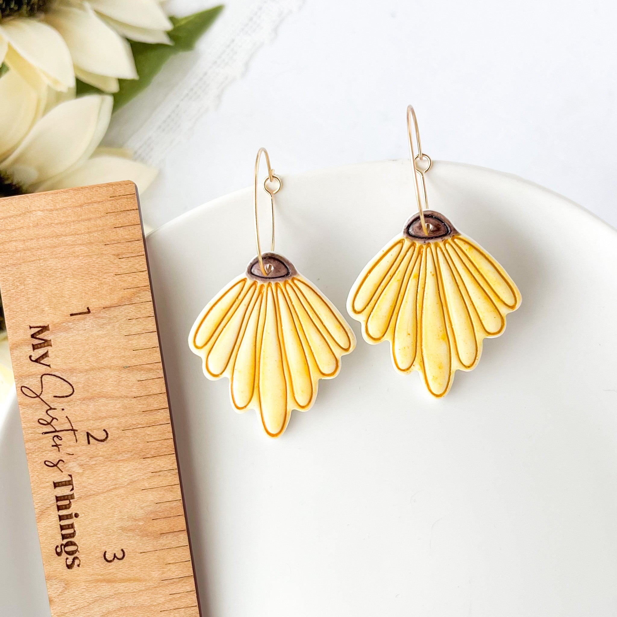 Yellow floral earrings on a white surface with a wooden ruler and flowers in the background.
