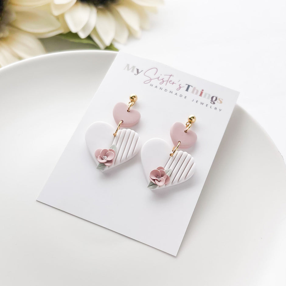 A pair of pink and white heart-shaped earrings with pink flowers and gold accents, displayed on a white plate with a floral decoration in the background.