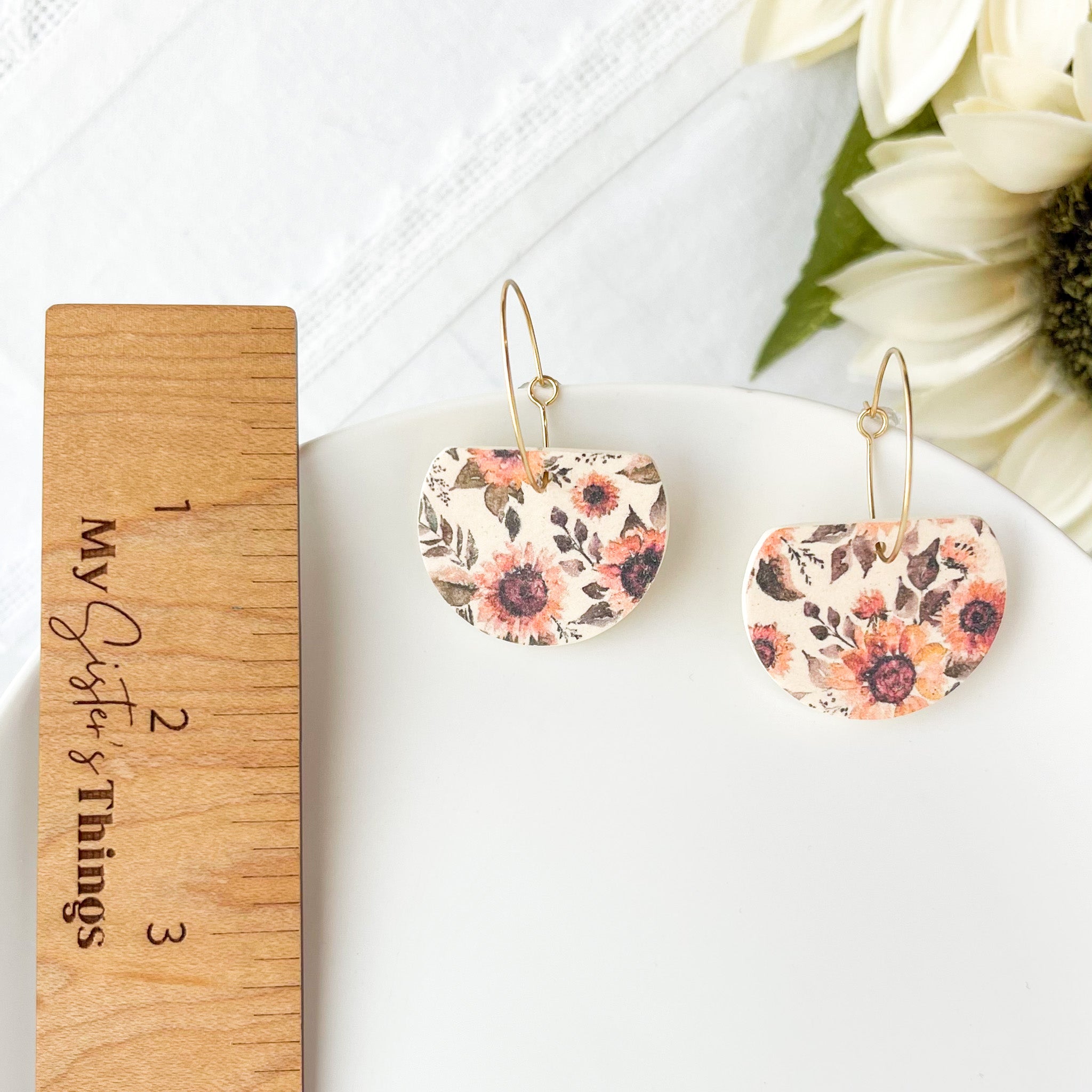 Floral earrings on a white surface with a wooden ruler and flowers in the background