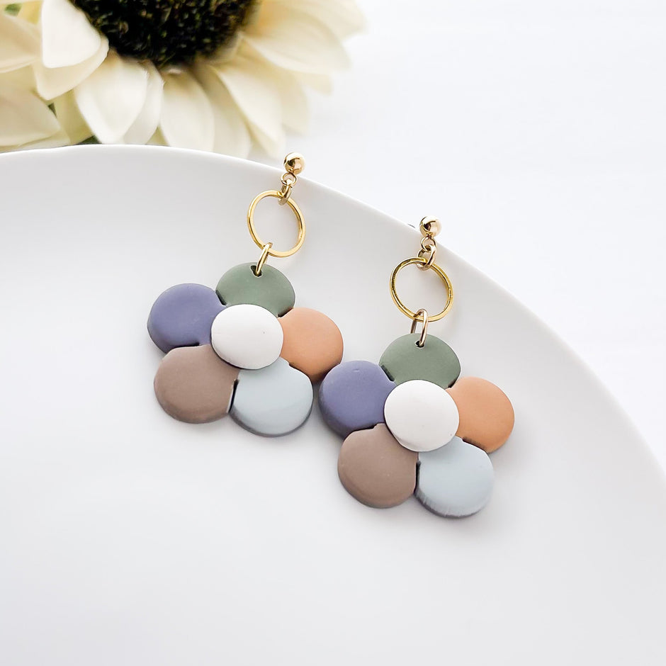 A pair of earrings featuring a geometric daisy flower pattern in shades of blue, green, and white, with gold accents, displayed on a white plate with a white background.