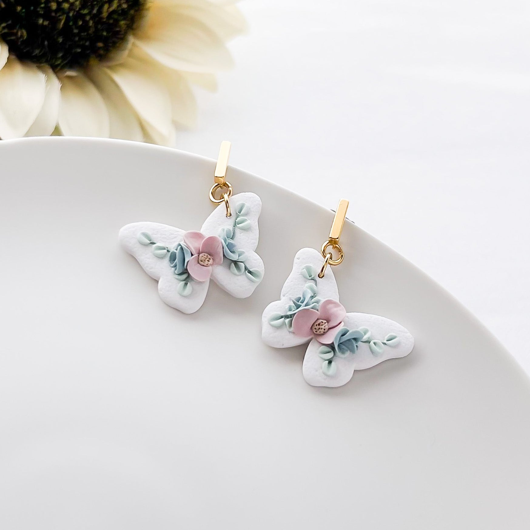 A pair of white and pink floral clip-on earrings with a gold-colored metal clip, displayed on a white plate with a sunflower in the background.