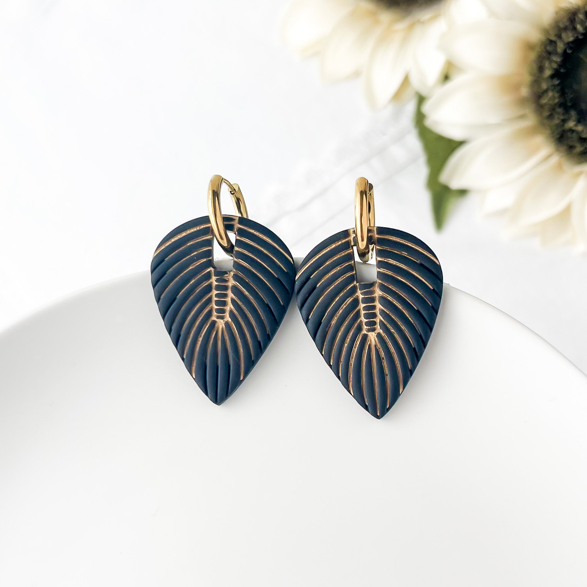 Pair of black leaf-shaped earrings with gold accents on a white surface with a sunflower in the background.