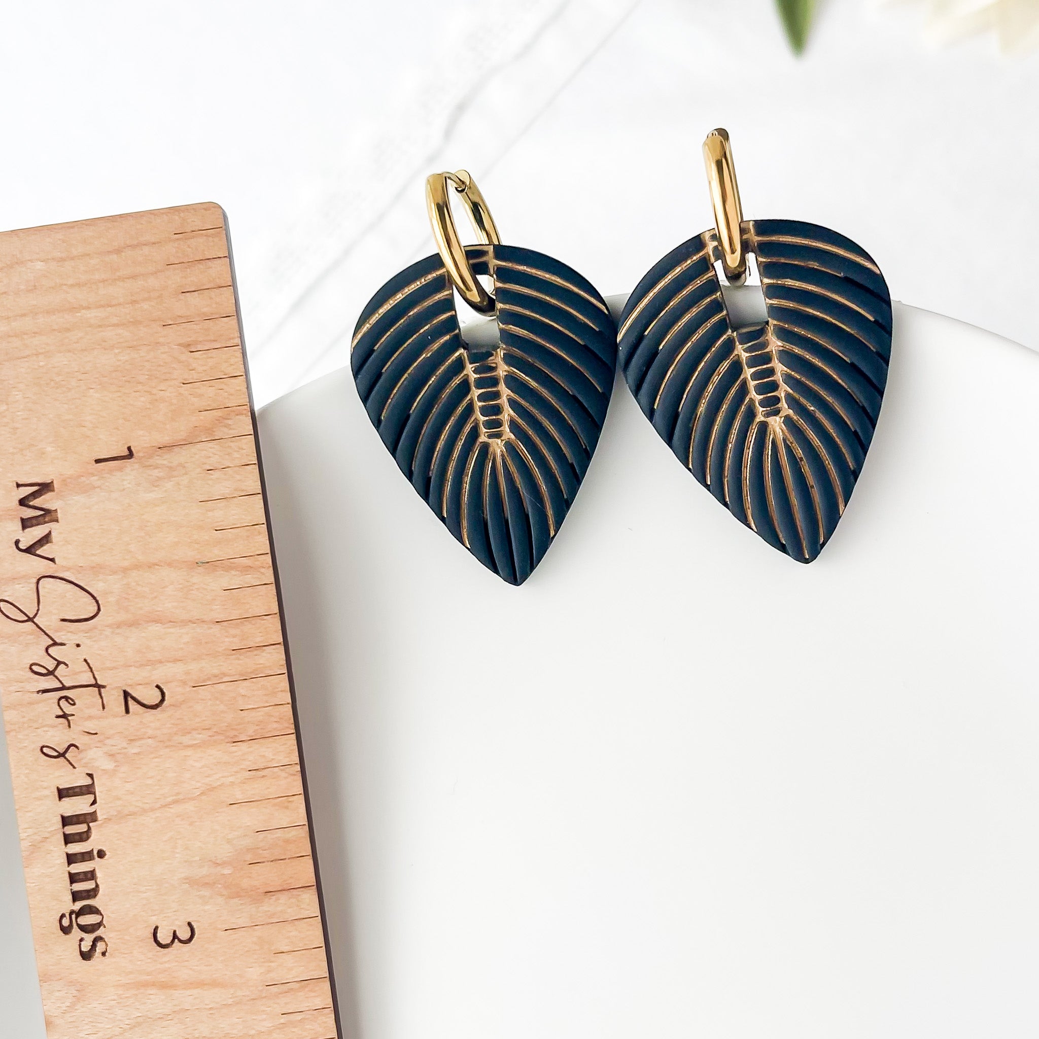 Pair of black leaf-shaped earrings with gold detailing on a white surface next to a wooden ruler.