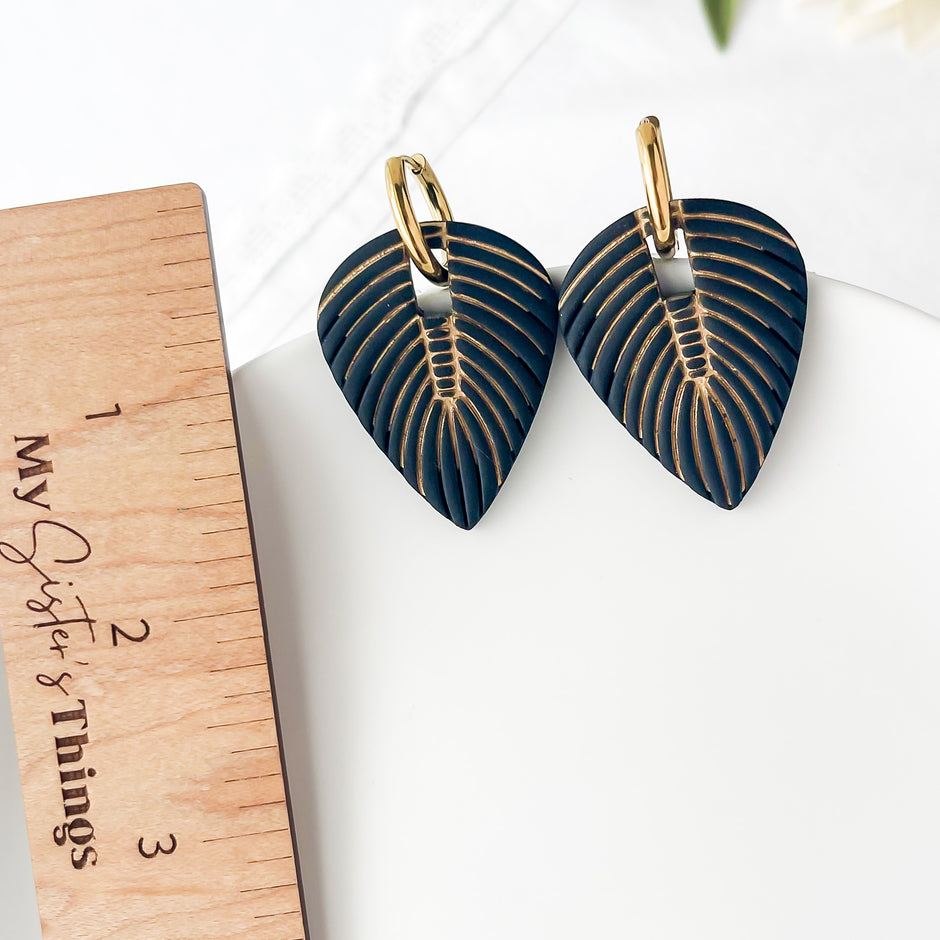 Pair of black leaf-shaped earrings with gold detailing on a white surface next to a wooden ruler.