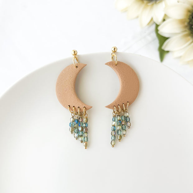 Crescent moon-shaped earrings with bead drops on a white plate with flowers in the background