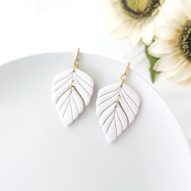 White leaf-shaped earrings on a white surface with sunflowers in the background
