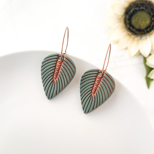 Green leaf-shaped earrings with copper wire detailing on a white surface with a sunflower in the background.