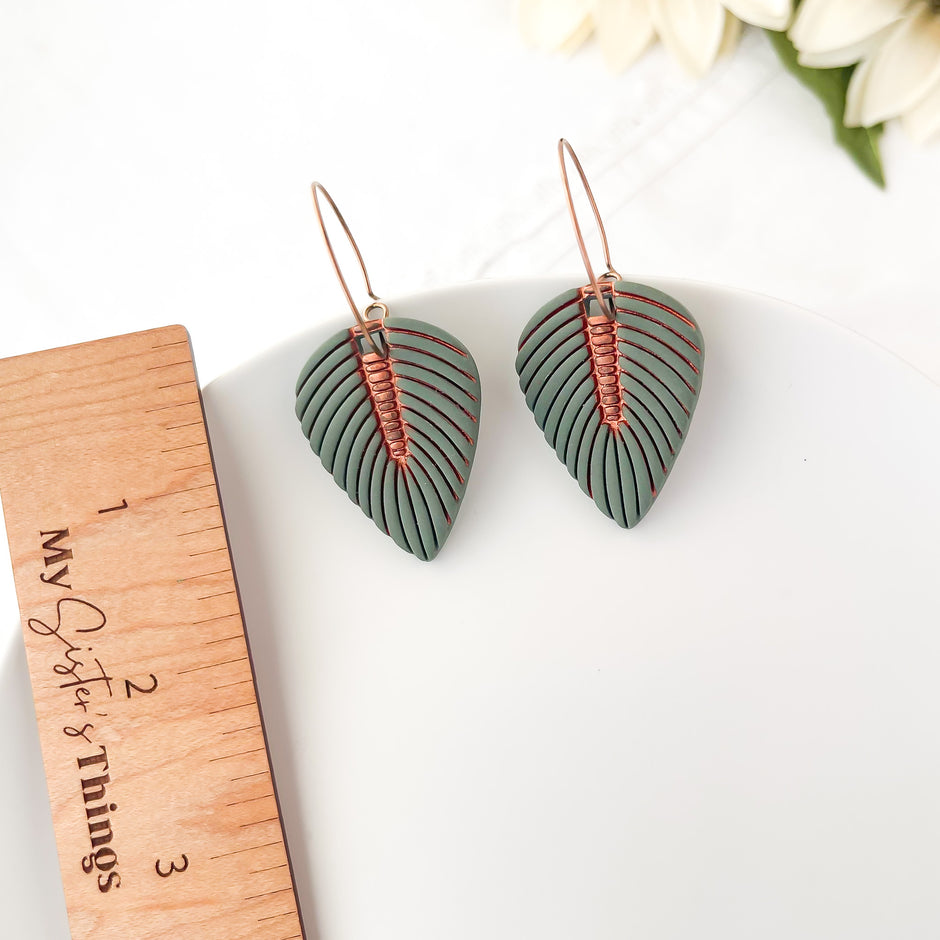 Pair of green leaf-shaped earrings with copper wire detailing on a white surface with a wooden ruler.