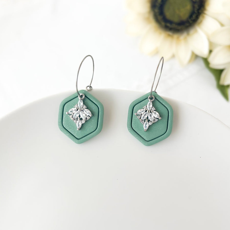 Mint green hexagonal earrings with silver accents on a white surface with flowers in the background