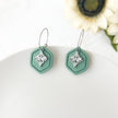 Mint green hexagonal earrings with silver accents on a white surface with flowers in the background