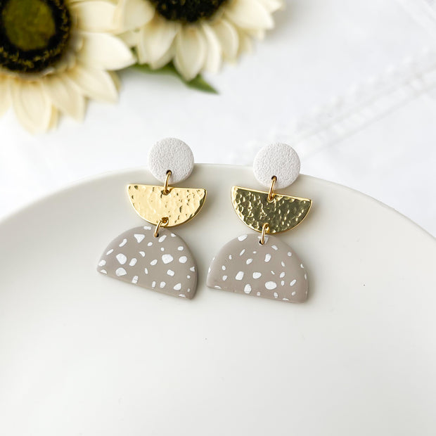 Pair of earrings with gold and taupe half-circle designs on a white surface with sunflowers in the background.