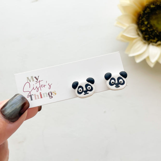 Panda face stud earrings held by a hand with a 'My Sister's Things' card and a sunflower in the background.