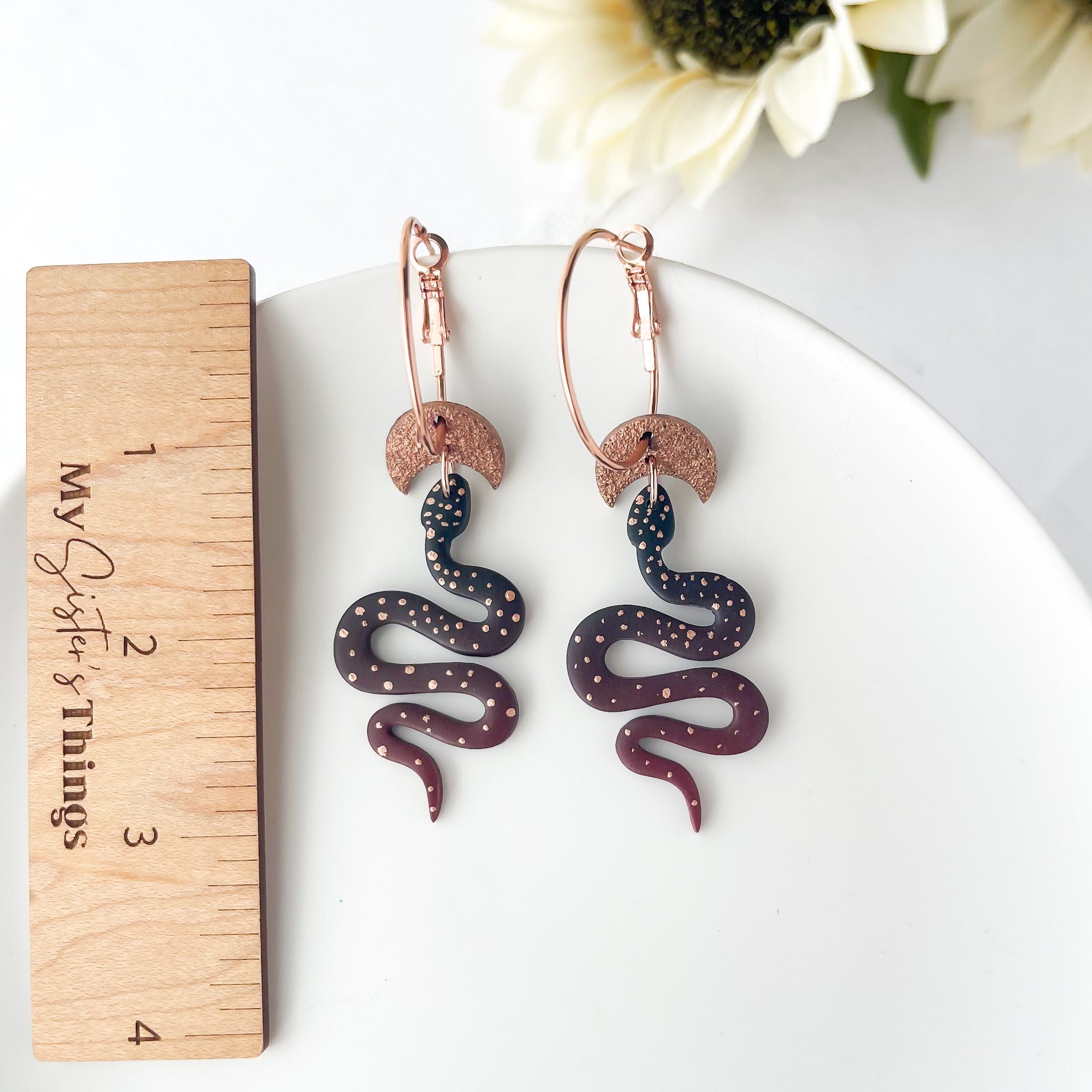 Snake-shaped earrings on a white plate with a wooden ruler for scale, against a white background with sunflowers.