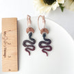 Snake-shaped earrings on a white plate with a wooden ruler for scale, against a white background with sunflowers.