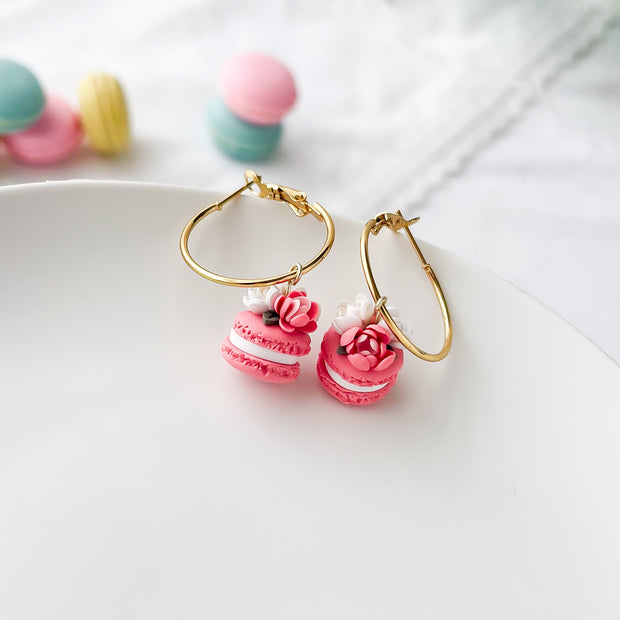 Gold hoop earrings with pink macaron-shaped charms on a white surface.
