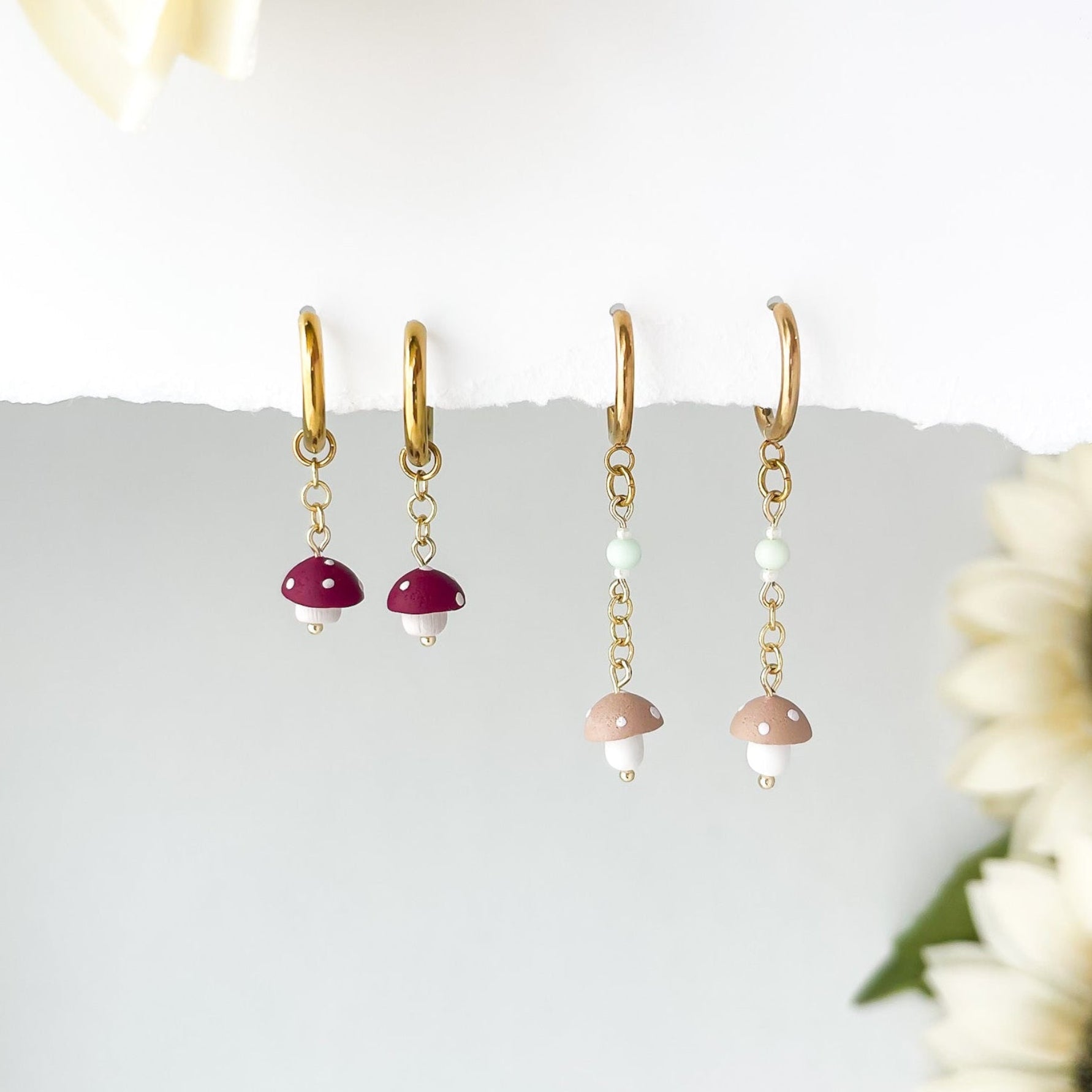 2 pair of tiny dangling mushroom earrings,  red and tan mushrooms hanging from gold chain  earring, with sunflowers in the background. 