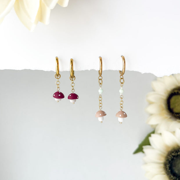 2 pair of tiny dangling mushroom earrings,  red and tan mushrooms hanging from gold chain  earring, with sunflowers in the background. 