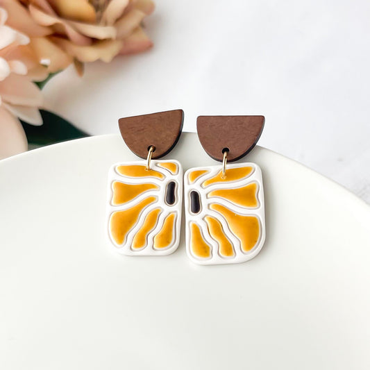 A pair of earrings with a retro sunflower design, featuring yellow and brown colors, displayed on a white plate with a floral background.