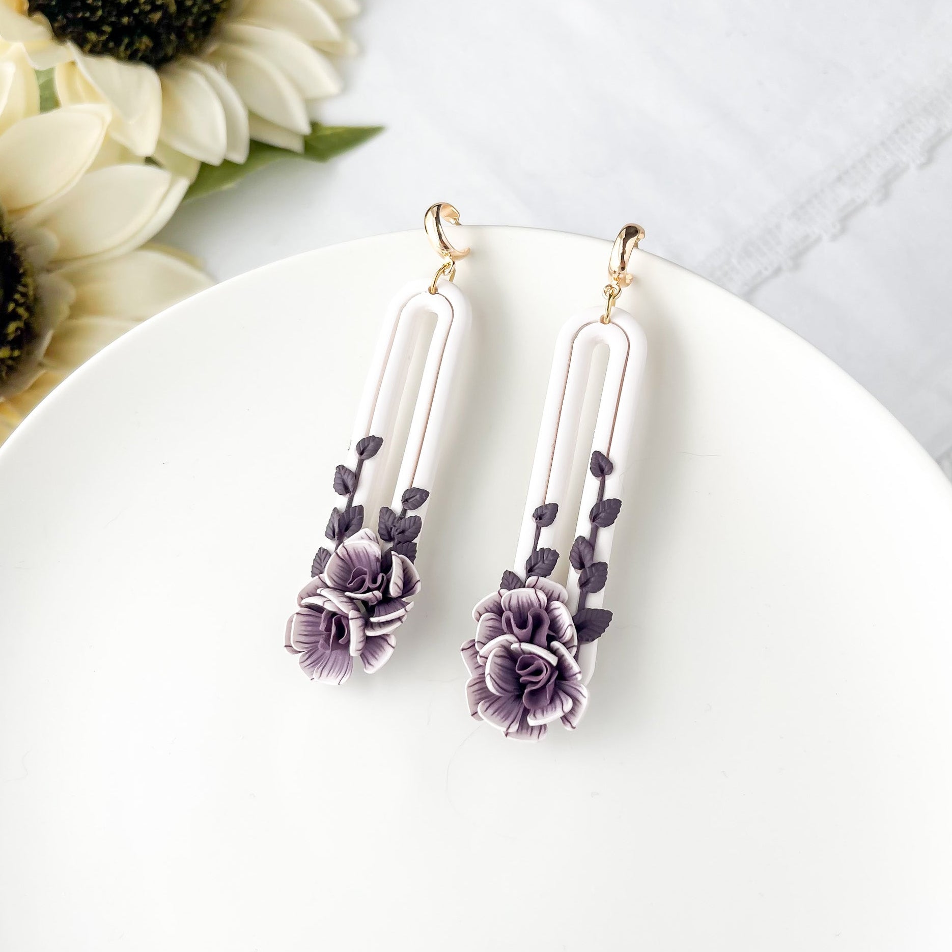 A pair of oval purple flower earrings with dangle design, placed on a white plate with a sunflower in the background.