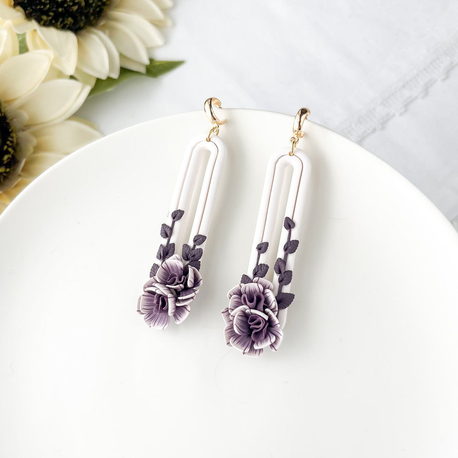 A pair of oval purple flower earrings with dangle design, placed on a white plate with a sunflower in the background.