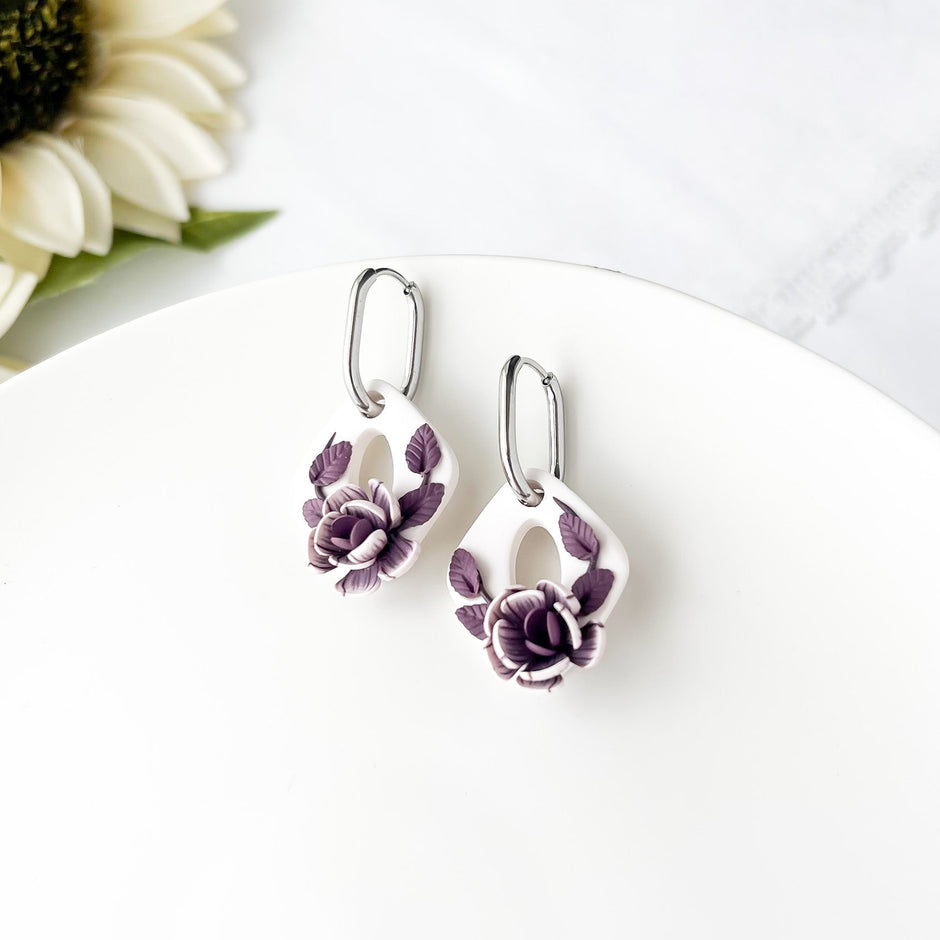A pair of purple floral earrings with a dangle design, placed on a white surface with a sunflower in the background.