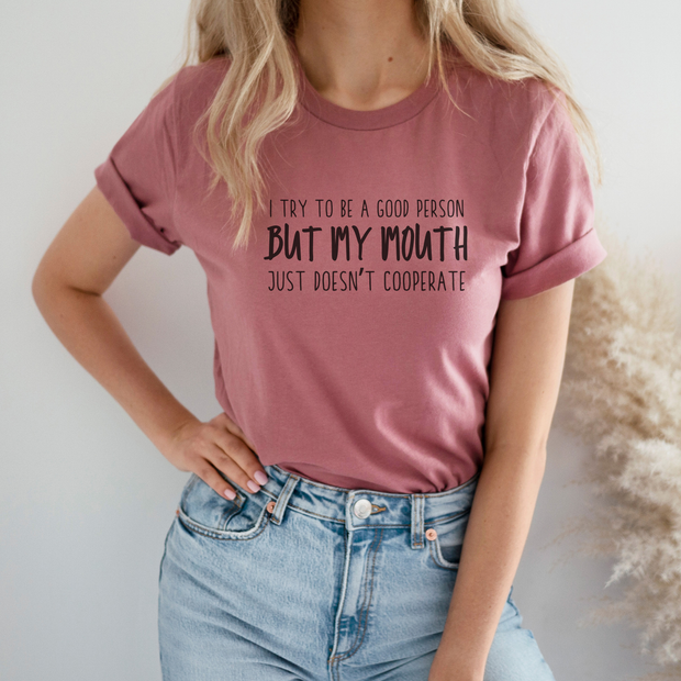 Person wearing a pink t-shirt with humorous text "I Try To Be A Good Person But My Mouth Just Doesn't Cooperate" on a plain background