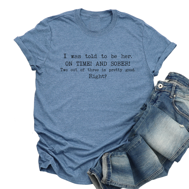 Blue t-shirt with humorous text "I Was Told to Be Here! On Time! And Sober! Two Out of Three Is Pretty Good. Right? Denim pants on a white background