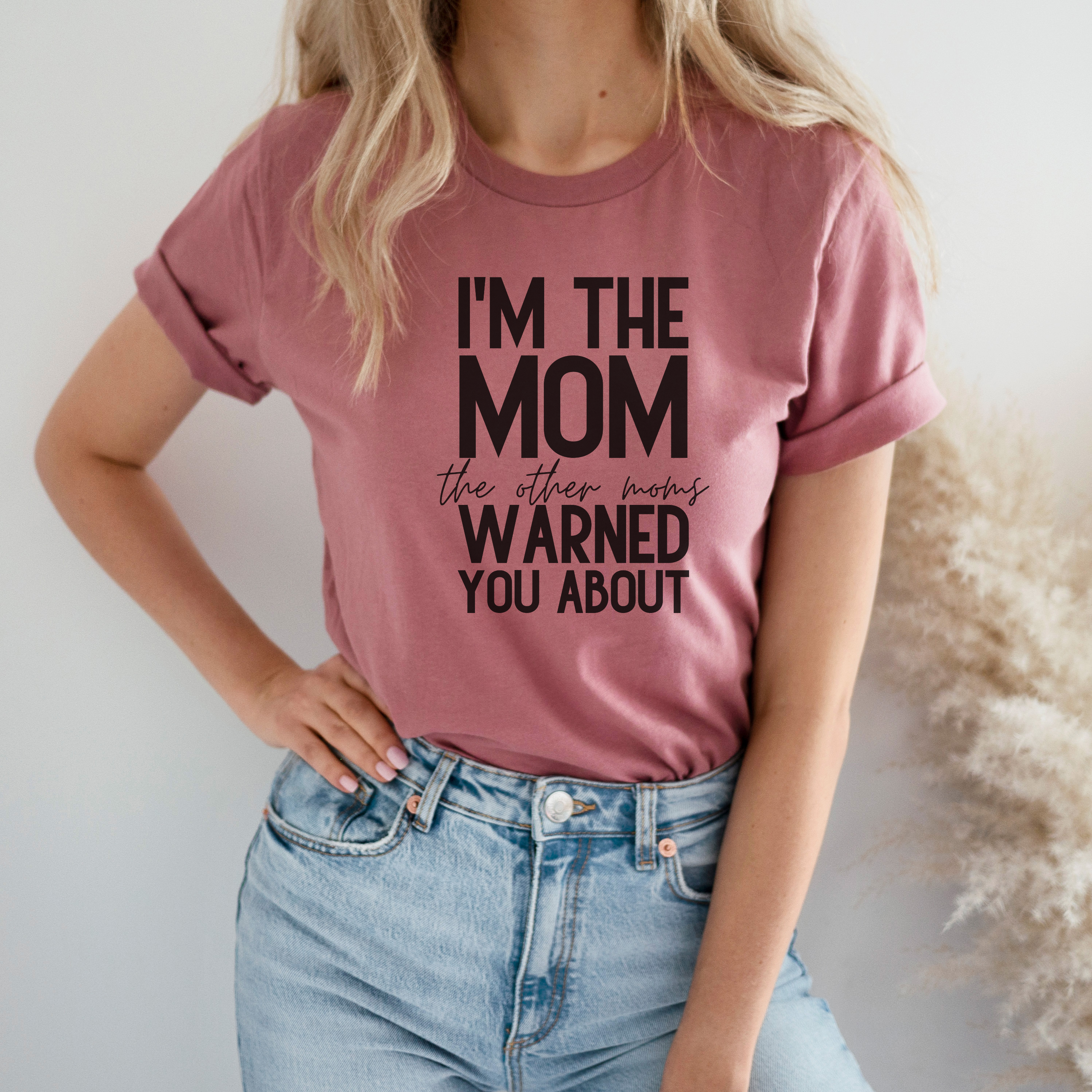 A woman wearing a pink t-shirt with the text 'I'm THE MOM THE OTHER MOM'S WARNED YOU ABOUT' printed on the front, paired with blue denim jeans.