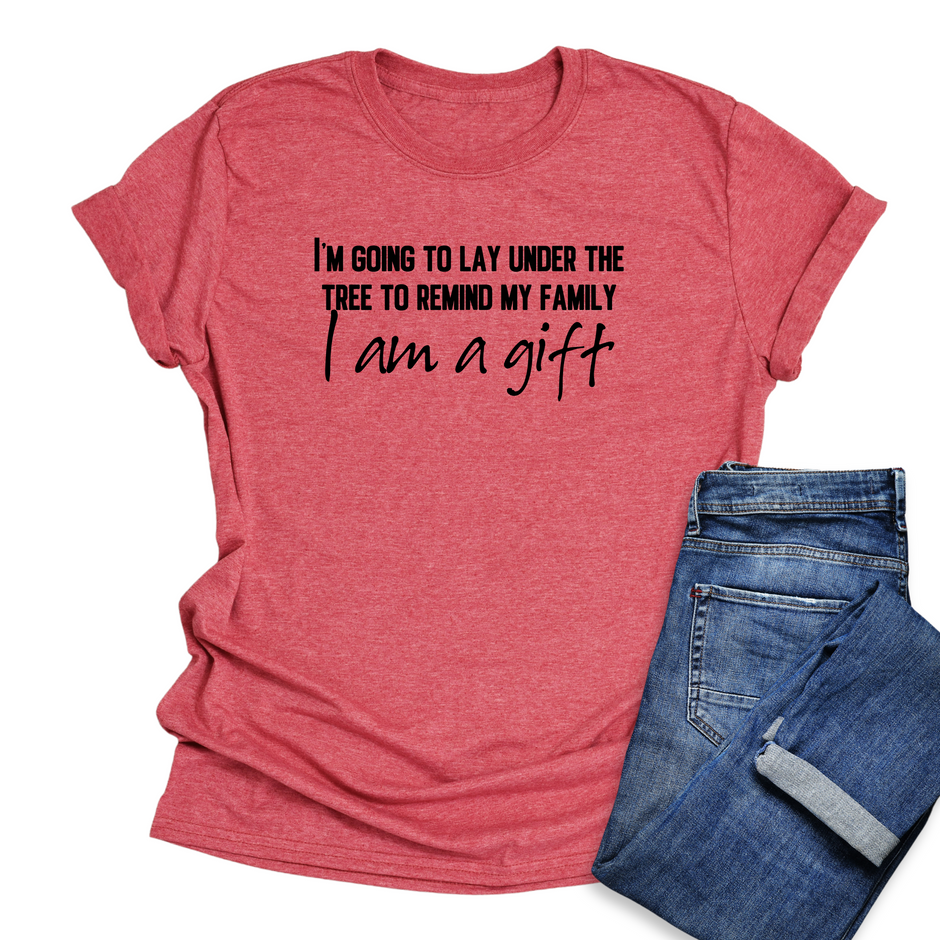 Red t-shirt with humorous text "I'm Going To Lay Under The Tree To Remind My Family I Am A Gift" with blue jeans on a white background