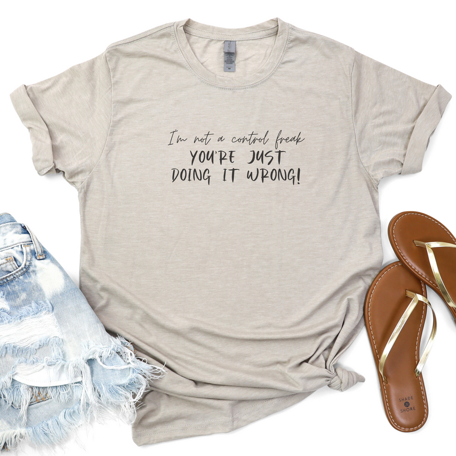 A silver unisex t-shirt with the text 'I'm Not A Control Freak You're Just Doing It Wrong!" printed on it, paired with blue denim shorts and flip flops. 
