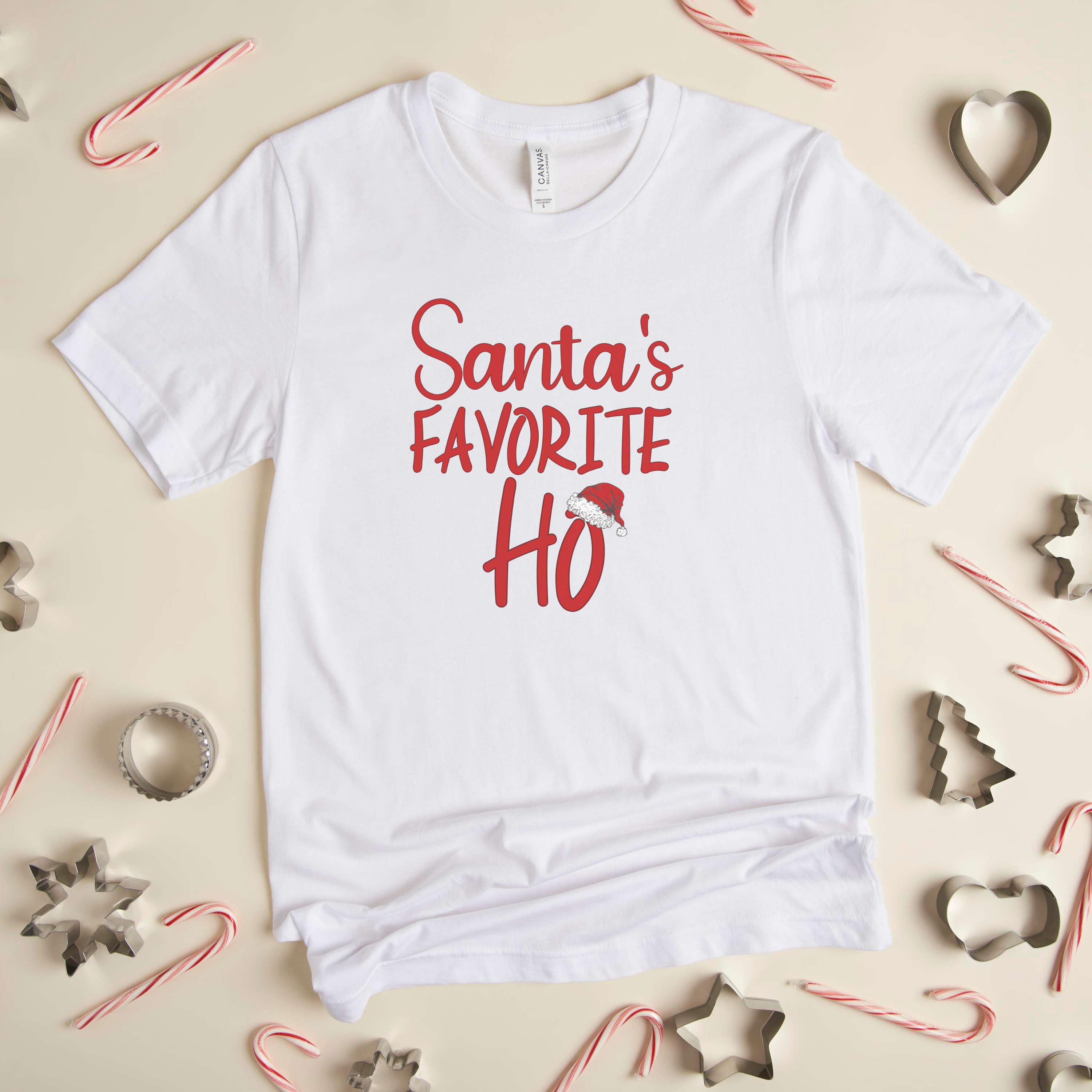 A white crew neck t-shirt with the text 'Santa's Favorite Ho' printed in red and green colors, displayed on a surface with candy canes and stars around it.