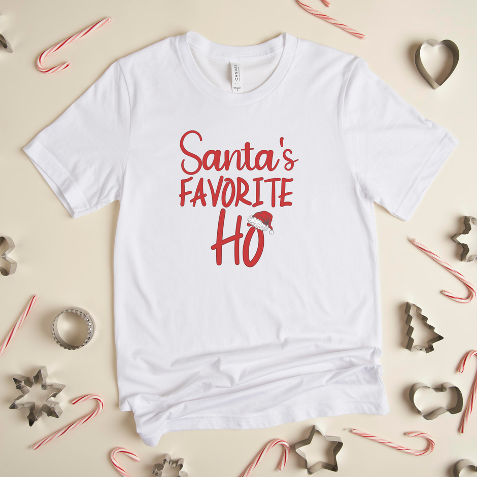 A white crew neck t-shirt with the text 'Santa's Favorite Ho' printed in red and green colors, displayed on a surface with candy canes and stars around it.
