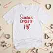 A white crew neck t-shirt with the text 'Santa's Favorite Ho' printed in red and green colors, displayed on a surface with candy canes and stars around it.