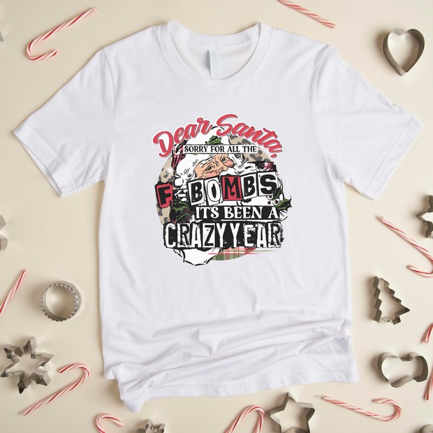White t-shirt with Christmas-themed text and graphics on a beige background with holiday decorations.