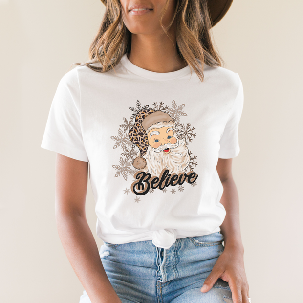 Person wearing a white t-shirt with a graphic of Santa Claus and the word 'Believe' on a plain background