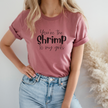 A woman wearing a pink t-shirt with the text 'You're The Shrimp To My Grits' printed on the front, paired with blue denim jeans.