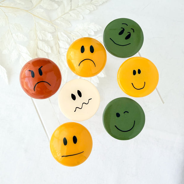 Round polymer clay fridge magnets with mixed expressions in terracotta, mustard, cream, and olive green.